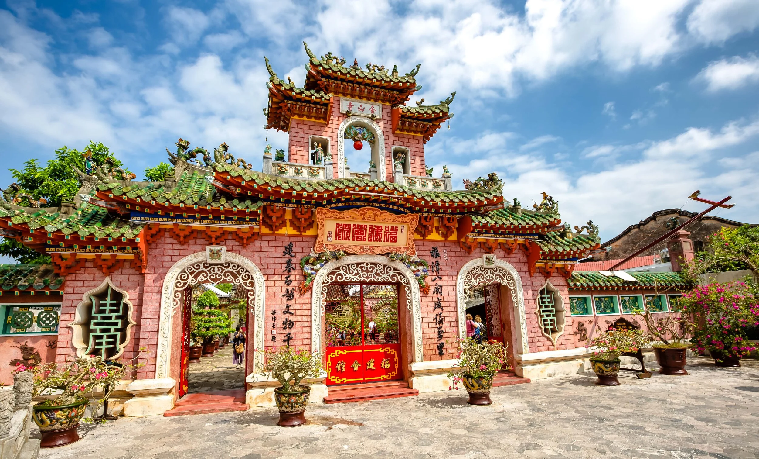 Colorful Chinese temple entrance with red walls, green tiled roofs, and ornate carvings under a blue sky with clouds.