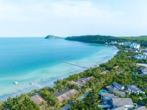 Aerial view of a tropical seaside town with palm-lined beach, turquoise water, and hillside hotels along the coast.