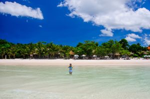 Child standing in clear turquoise water at a tropical beach with palm trees and thatched huts along the shore under a blue sky.