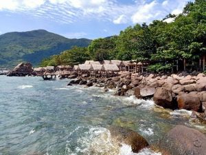 Thatched-roof huts line a rocky shoreline with clear blue water and forested hills in the background.