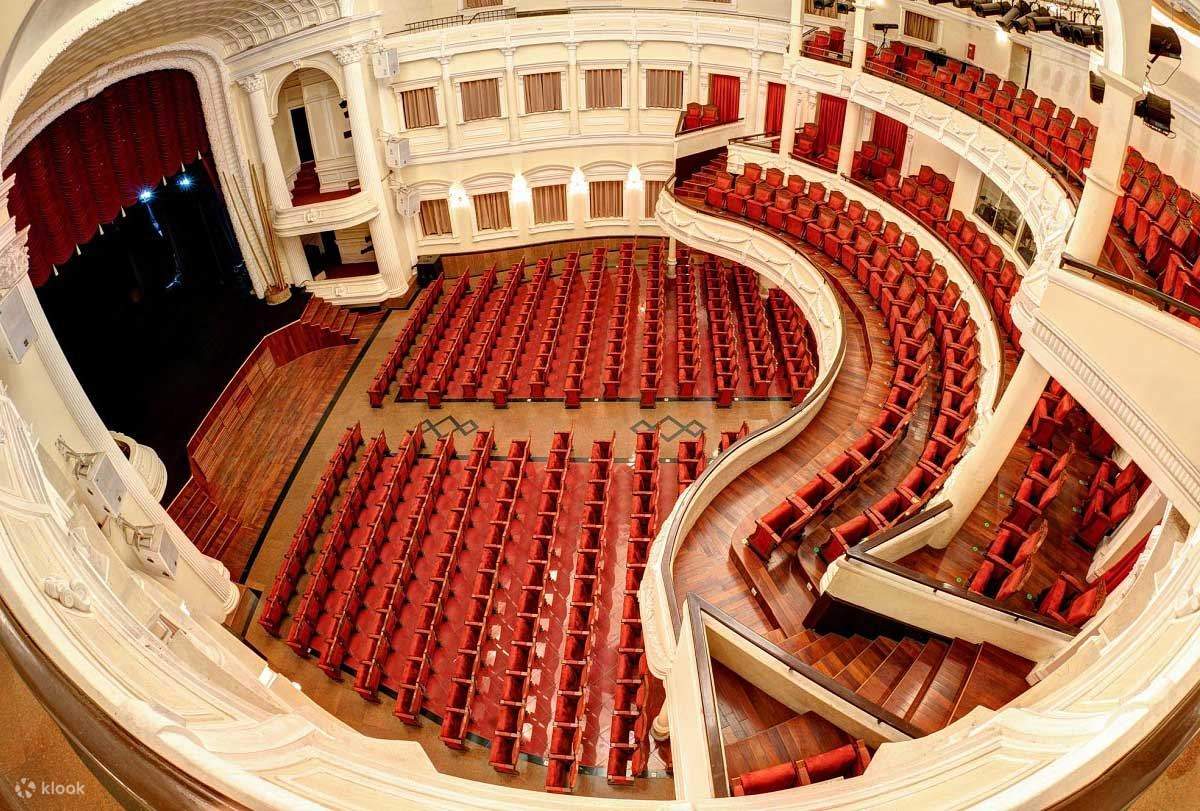 An empty classical theater with curved white balconies and rows of red upholstered seats viewed from above.