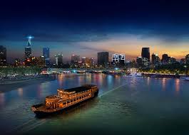 Dusk over a city skyline along a river, with a lit ferry slowly gliding across the foreground water.
