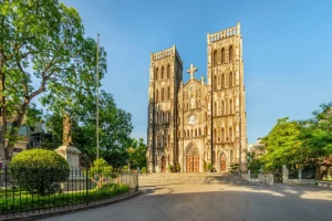 Gothic cathedral with twin towers and a central rose window, set in a quiet park with a statue and greenery to the left.