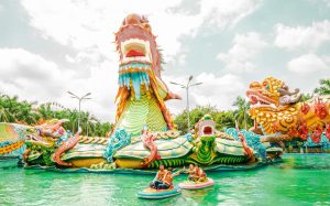 Colorful dragon-themed water ride with riders in bumper boats on a green pool, under a bright cloudy sky