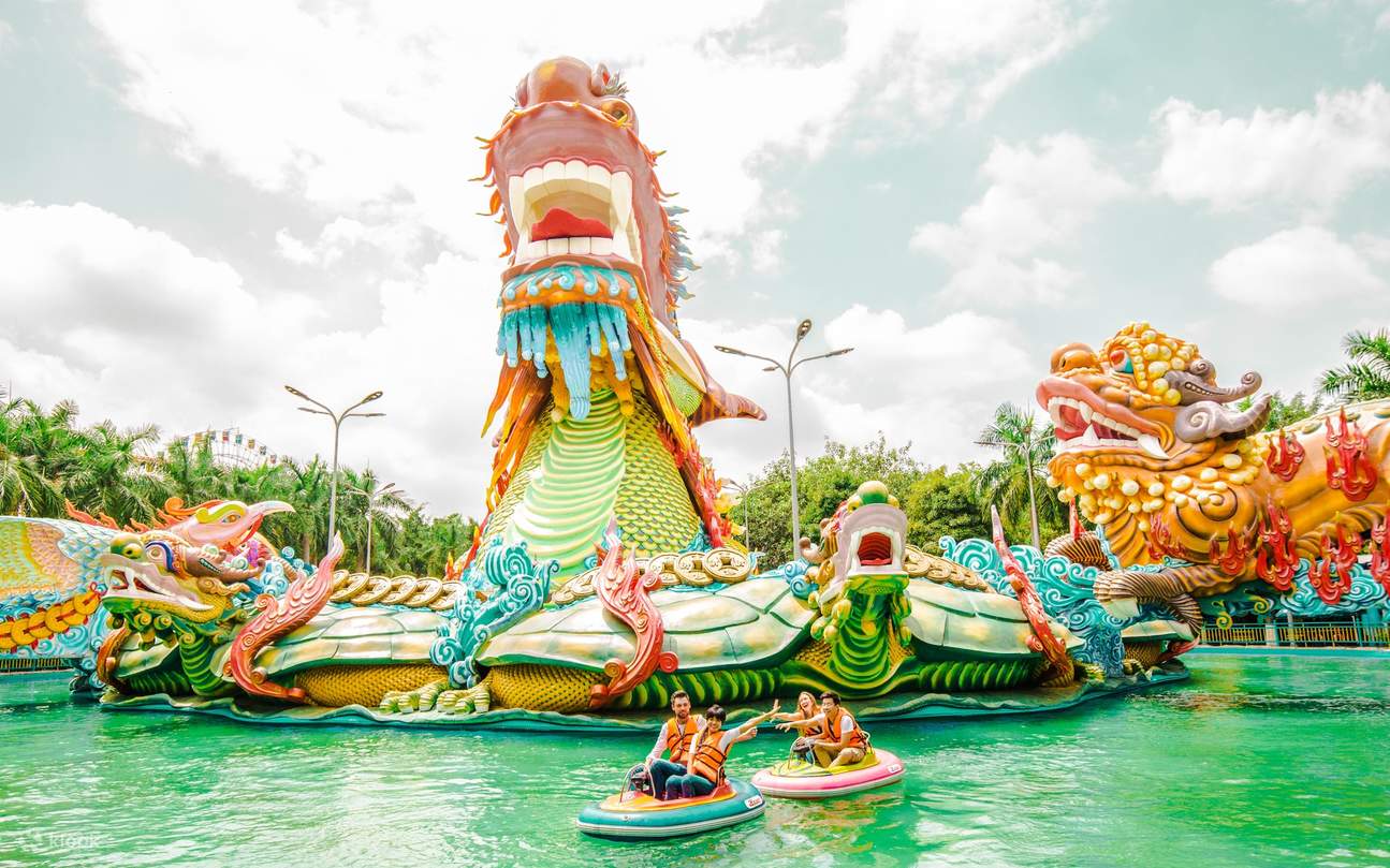 Colorful dragon-themed water ride with riders in bumper boats on a green pool, under a bright cloudy sky