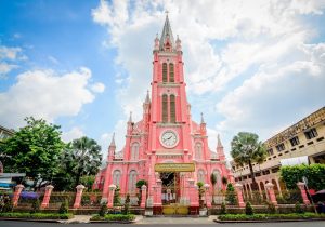 Front view of a tall pastel pink church with a clock on the façade and a pointed central steeple, set against a blue sky with palm trees and a decorative fence nearby.