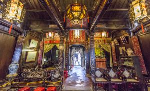 Intricately carved wooden interior of a Chinese hall with hanging lanterns, antique furniture, and an altar at the far end leading to a courtyard.