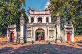 Ornate stone gate with a central arch and side doors, set in a park with trees and a pathway behind the arch.