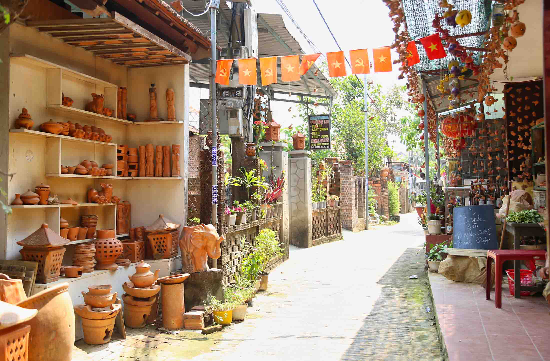 Narrow street market with shelves of terracotta pots on the left and orange flags overhead.