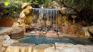Rock waterfall cascading into a turquoise pool, surrounded by large stones in a garden setting.