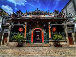 Front view of an ornate Asian temple entrance with red doors, hanging lanterns, and potted plants on a stone courtyard under a blue sky.
