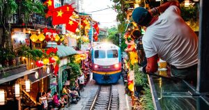 Blue and white train on a narrow street flanked by colorful lanterns, flags, and people watching from both sides.