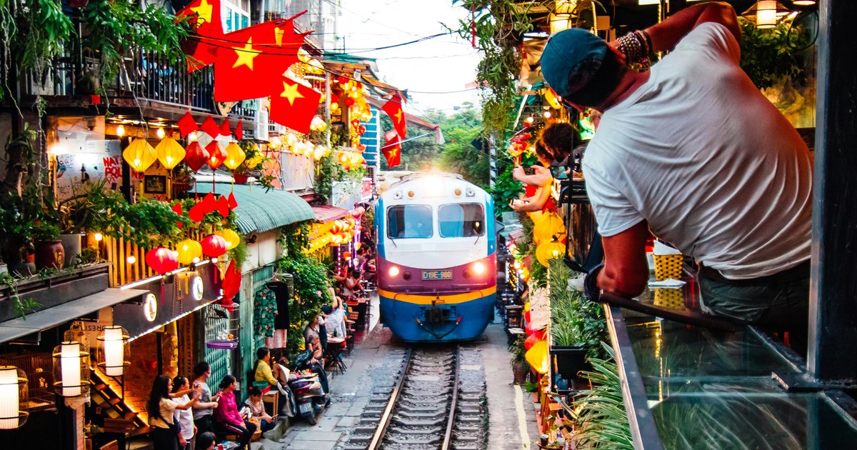 Blue and white train on a narrow street flanked by colorful lanterns, flags, and people watching from both sides.