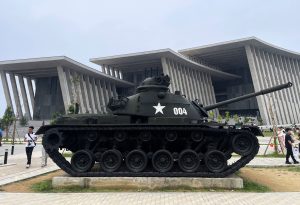 A large military tank on display in a plaza with a modern, columned building in the background and pedestrians nearby.