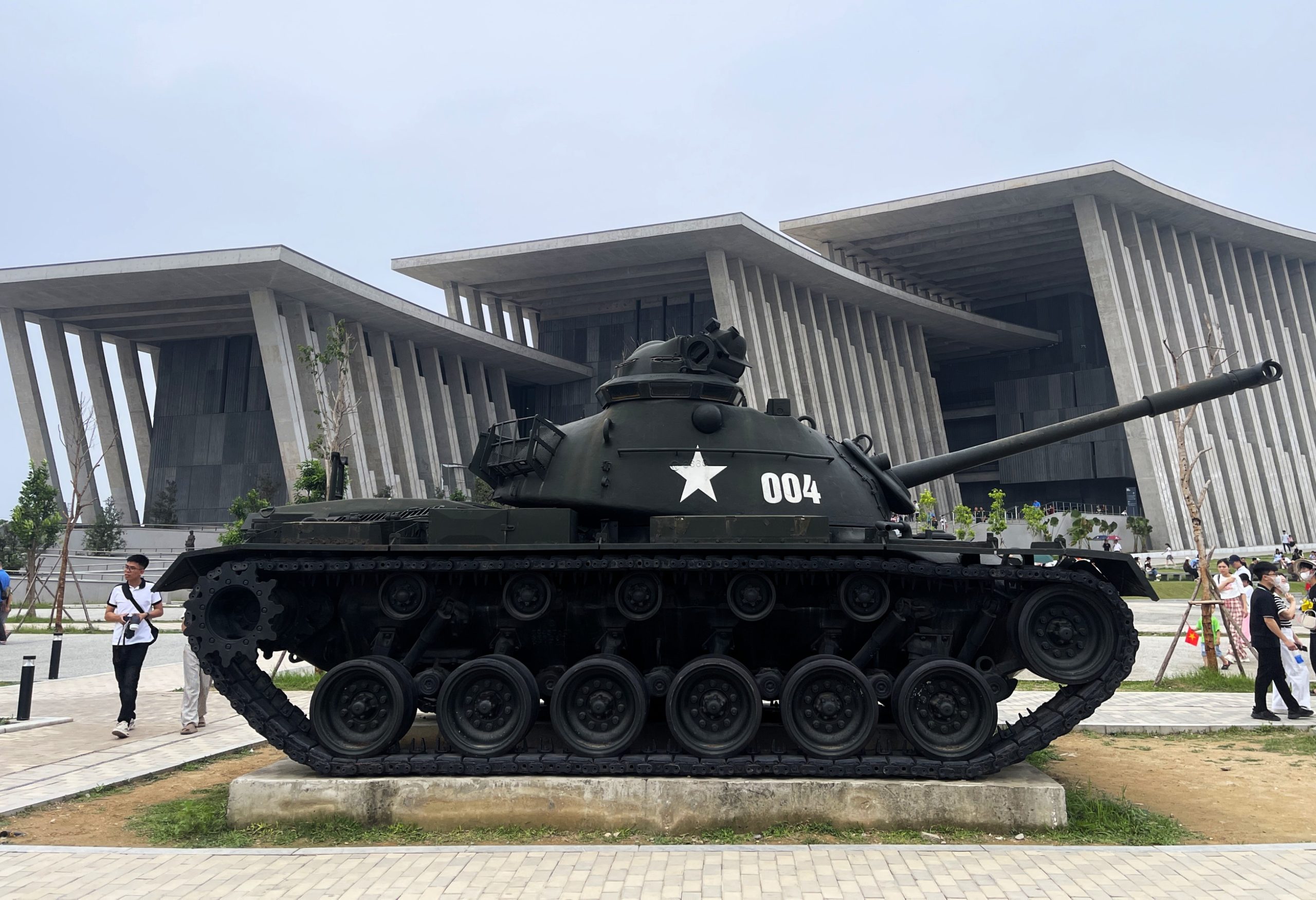 A large military tank on display in a plaza with a modern, columned building in the background and pedestrians nearby.