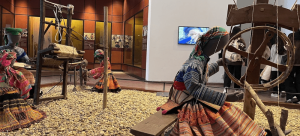 Museum exhibit showing traditional weaving with life-size mannequins in colorful dresses and a wooden loom on a pebble floor, with visitors in the background.