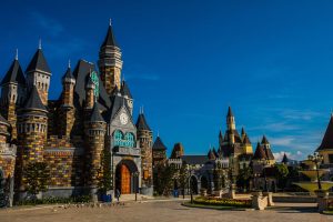 Fantasy castle complex with stone towers and a wooden gate under a clear blue sky at a theme park plaza.