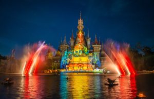 Colorful illuminated castle at night with water jets arcing over a reflecting lake.