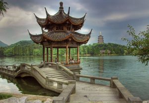 Traditional Chinese pavilion on a lake, connected by a curved stone bridge, with a distant pagoda and green forest.