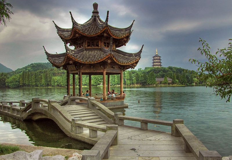 Traditional Chinese pavilion on a lake, connected by a curved stone bridge, with a distant pagoda and green forest.
