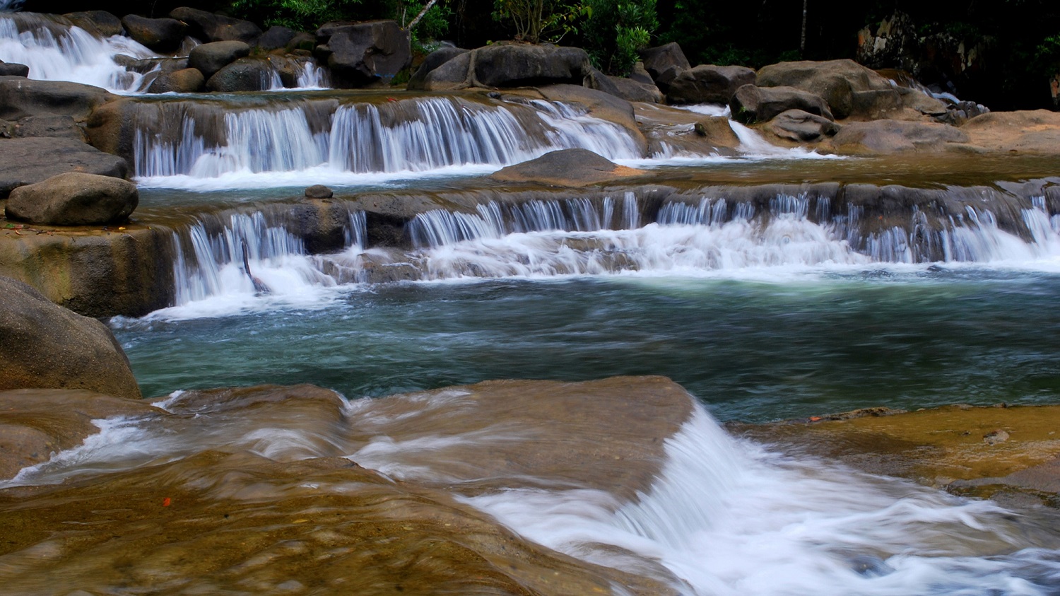 Tiered waterfall flowing over smooth brown rocks into a turquoise pool