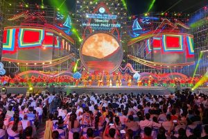Colorful festival stage with dancers performing in orange outfits, bright geometric decorations, and a large circular moon backdrop; audience seated and filming the show at night.