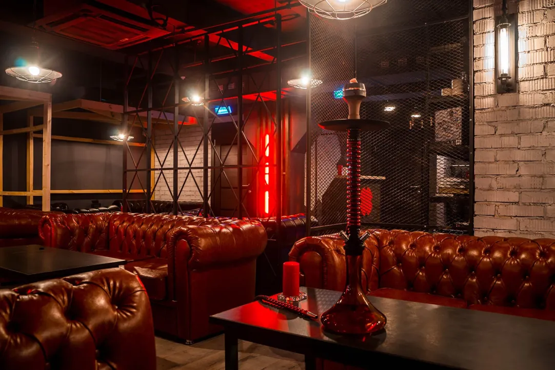 Hookah on a table in a dim lounge with tufted brown leather sofas and red ambient lighting.