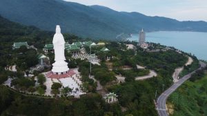 Aerial view of a hillside temple complex with a tall white statue, green-roofed buildings, and a multi-story pagoda by the coast.