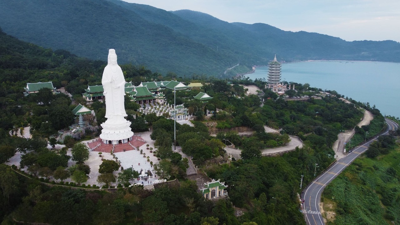 Aerial view of a hillside temple complex with a tall white statue, green-roofed buildings, and a multi-story pagoda by the coast.