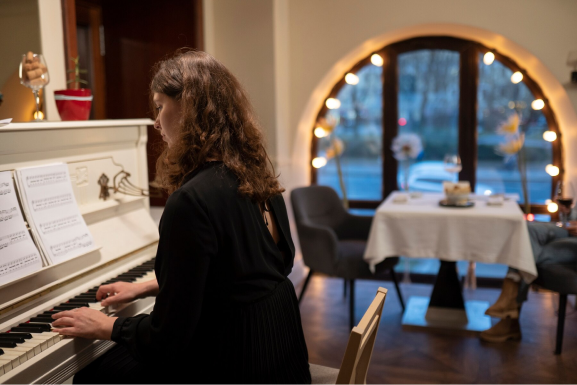 Woman with curly hair in black plays a white upright piano; a dining table and warm, glowing lights are in the background.