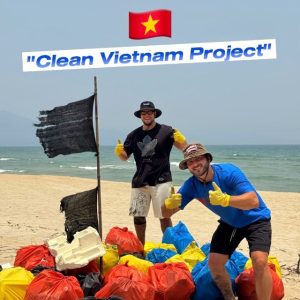 Two volunteers on a sunny beach give thumbs up beside colorful trash bags, under a 'Clean Vietnam Project' banner with a Vietnamese flag above them.