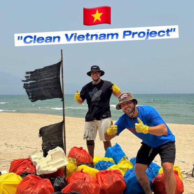 Two volunteers on a sunny beach give thumbs up beside colorful trash bags, under a 'Clean Vietnam Project' banner with a Vietnamese flag above them.