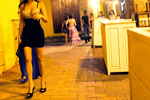 Woman in a black mini dress and heels walking down a warmly lit street, with people in the background.
