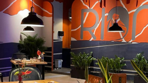 Restaurant dining area with a bold orange wall mural featuring large letters reading 'RIO', hanging pendant lights, wooden tables, and potted plants.