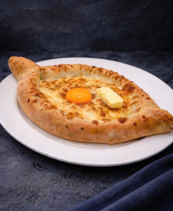Boat-shaped Georgian cheese bread (khachapuri) with a sunny-side-up egg and a butter pat on top, served on a white plate.