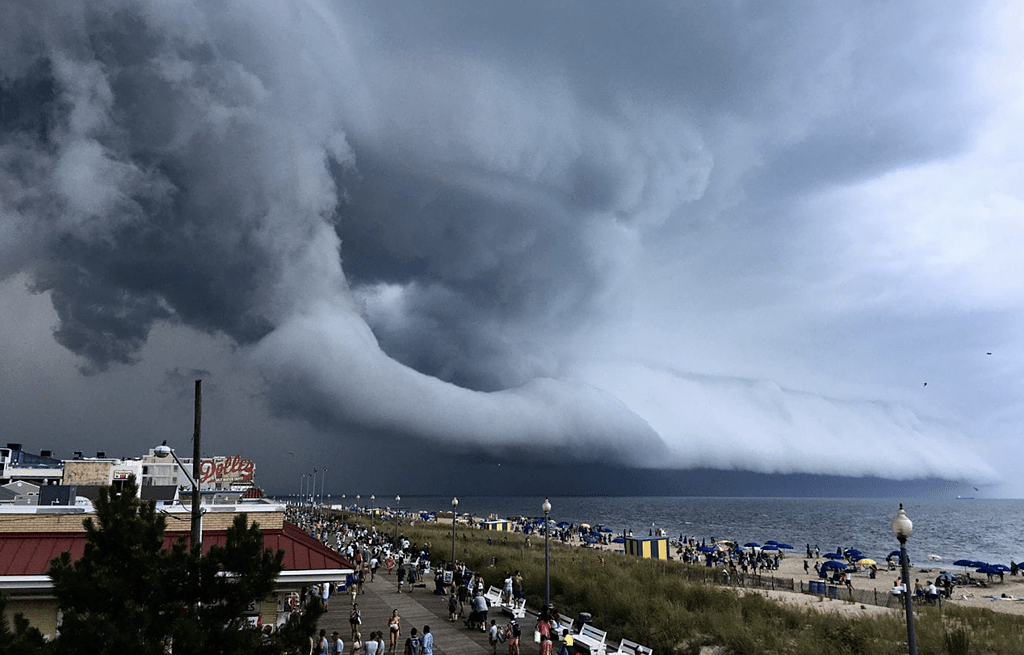 Gigantic shelf cloud rolling toward the beach over the lake as people walk along a crowded boardwalk and the shore.