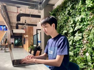Young man in a navy T‑shirt types on a laptop at a desk beside a lush vertical garden in a modern coworking space.