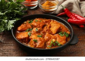 Beef stew chunks in a tomato-based sauce garnished with parsley in a cast-iron skillet on a wooden table, with red chilies nearby.