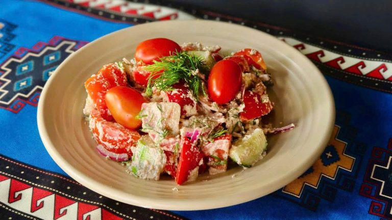 Beige plate of tomato, cucumber and red onion salad with feta and dill, on a blue patterned tablecloth.