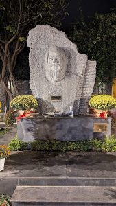 Stone bust sculpture of a bearded man on a monument, flanked by yellow chrysanthemums, in a nighttime garden setting.