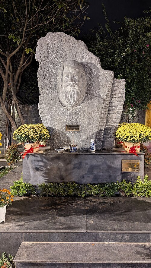 Stone bust sculpture of a bearded man on a monument, flanked by yellow chrysanthemums, in a nighttime garden setting.