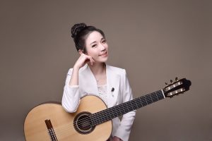 Young woman in a white blazer holding a classical guitar, resting her chin on her hand and smiling at the camera.