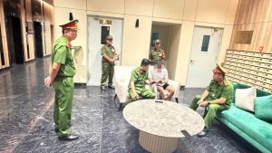 Guard officers in green uniforms stand and sit in a modern lobby with a round marble table and a teal sofa; a man in casual clothes sits on a beige chair between them.