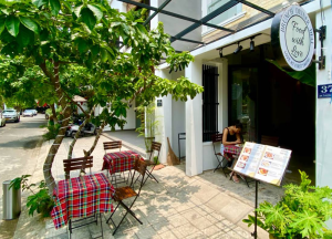 Outdoor cafe with red plaid tablecloths, wooden chairs, and leafy trees on a sunny sidewalk; a customer looks at a menu outside the restaurant.